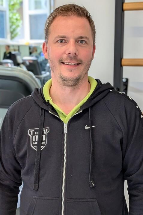 Mann l&auml;chelt in Sportkleidung vor Fitnessger&auml;ten in einem modernen Trainingsraum.