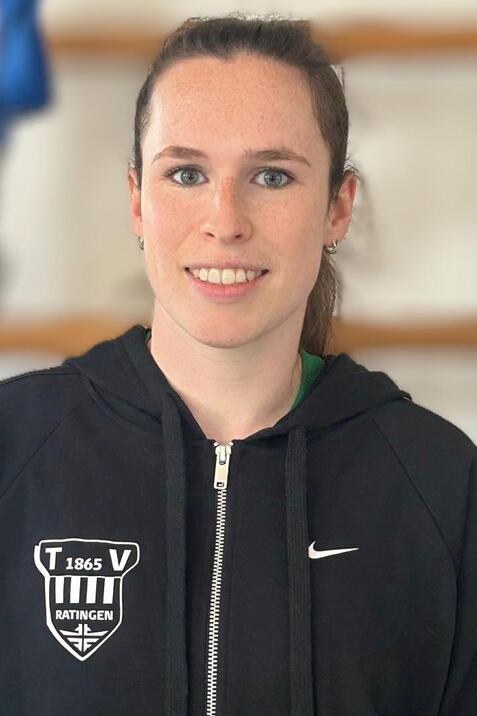 Portr&auml;t einer sportlichen Frau in schwarzer Jacke mit Logo, l&auml;chelnd vor einem neutralen Hintergrund.