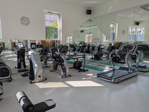 Fitnessraum mit verschiedenen Trainingsgeräten, Spiegelwand und einem Wandux mit Uhr. Tageslicht strömt durch große Fenster.