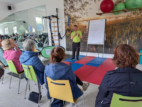 Mann in gr&uuml;ner Shirt erkl&auml;rt einer Gruppe von sitzenden Personen in einem Fitnessraum an einem Flipchart.