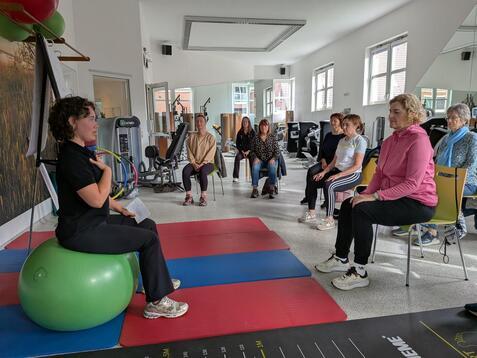 Eine Trainerin sitzt auf einem Gymnastikball und leitet eine Gruppe von Frauen in einem Fitnessraum an.