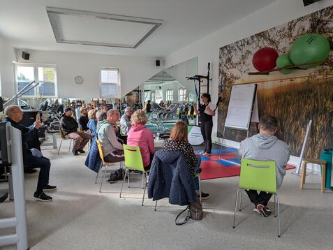 Gruppe &auml;lterer Menschen sitzt auf St&uuml;hlen in einem Fitnessraum; eine Trainerin erkl&auml;rt an einem Flipchart.