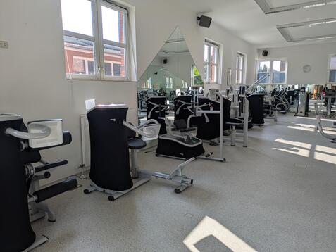 Innenansicht eines modernen Fitnessstudios mit mehreren Geräten und großen Fenstern.