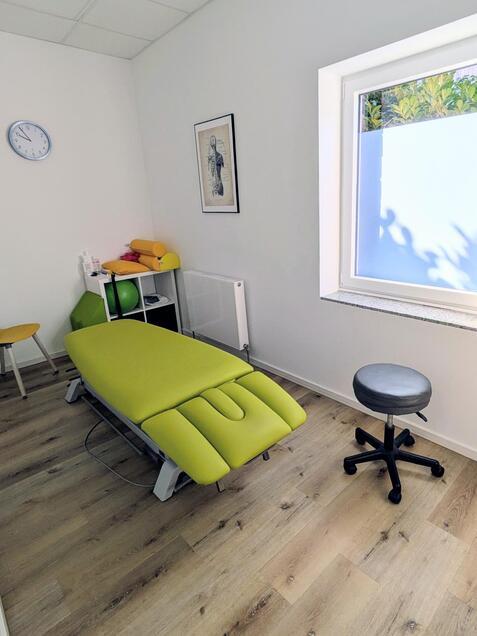Therapieraum mit grüner Liege, schwarzem Drehstuhl und Fenster, hell erleuchtet durch Tageslicht.