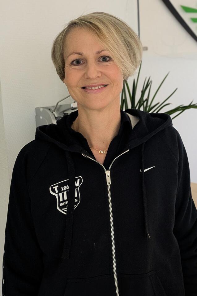 Frau mit kurzem, blonden Haar tr&auml;gt einen schwarzen Rei&szlig;verschluss-Hoodie und steht vor einer Wand mit dem Logo &bdquo;Ratingen&ldquo;.