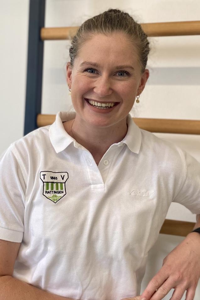 Portrait einer lächelnden Frau in einem weißen Poloshirt, die an einer Wand mit Holzsprossen steht.