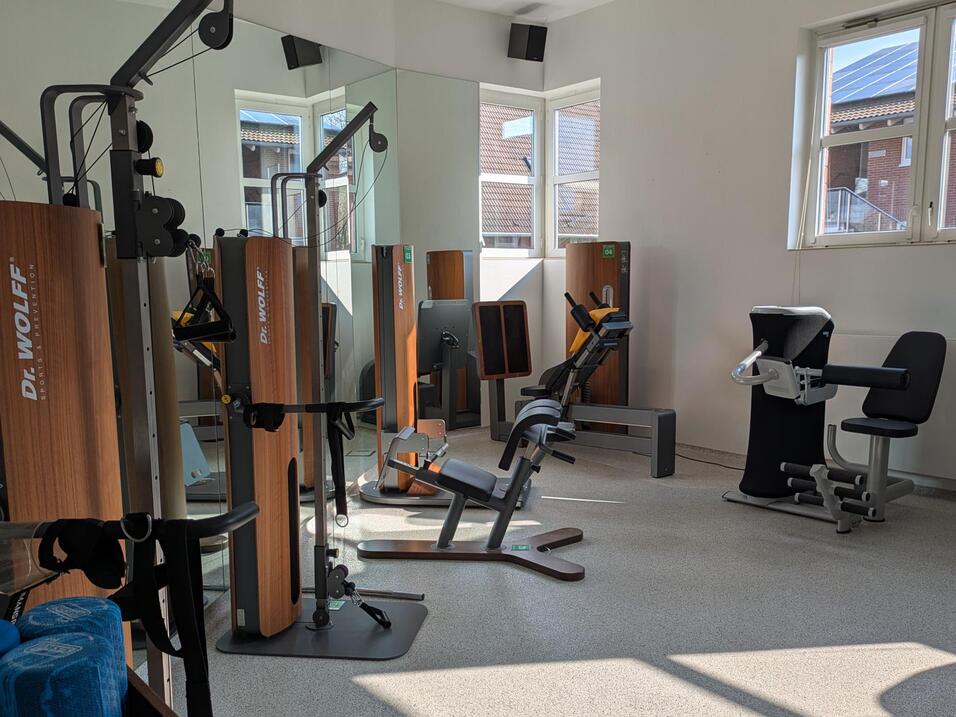 Fitnessraum mit verschiedenen Trainingsgeräten und Freihanteln, hell erleuchtet durch große Fenster.