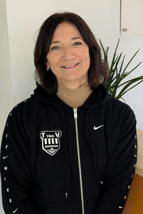 Frau in schwarzer Sportjacke mit Logo l&auml;chelt vor einer Empfangstheke mit Pflanzen im Hintergrund.