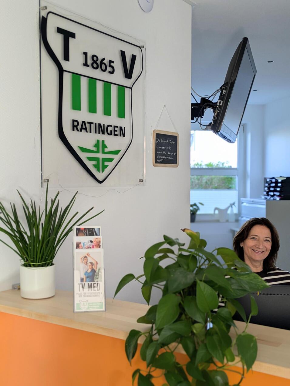 Empfangsbereich mit einem Vereinswappen, einer Pflanze, einer Informationsbrosch&uuml;re und einer l&auml;chelnden Frau am Empfangstresen.