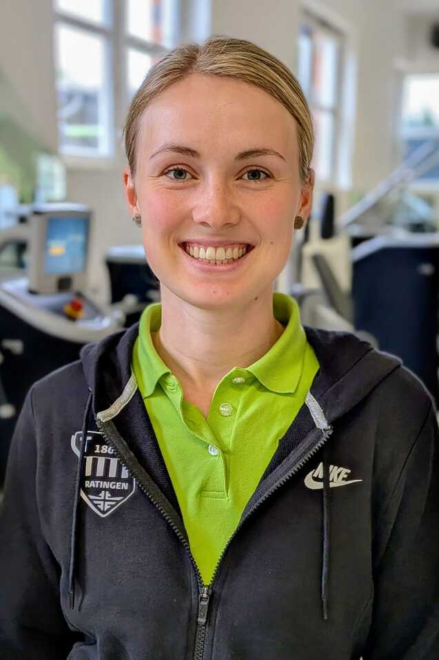 Lächelnde Frau mit kurzen blonden Haaren in Sportkleidung, steht in einem Fitnessstudio mit Trainingsgeräten im Hintergrund.