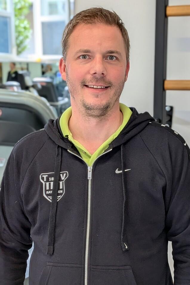 Mann l&auml;chelt in Sportkleidung vor Fitnessger&auml;ten in einem modernen Trainingsraum.