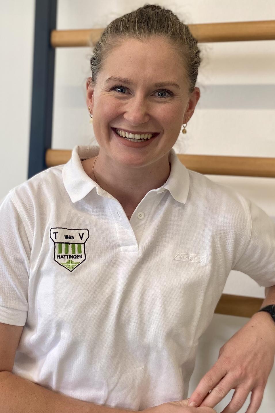 Portrait einer l&auml;chelnden Frau in einem wei&szlig;en Poloshirt, die an einer Wand mit Holzsprossen steht.