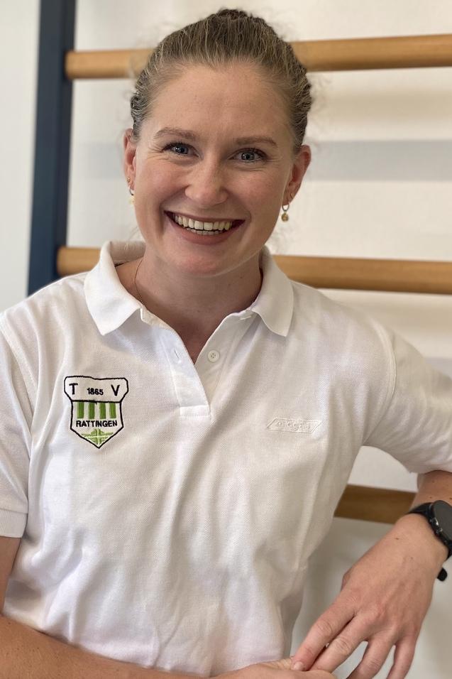 Portrait einer l&auml;chelnden Frau in einem wei&szlig;en Poloshirt, die an einer Wand mit Holzsprossen steht.