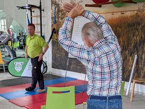 &Auml;lterer Mann dehnt sich mit erhobenen Armen in einem Fitnessraum, w&auml;hrend ein Trainer im Hintergrund zusieht.
