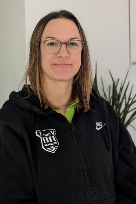 Portr&auml;t einer Frau mit Brille, die einen schwarzen Hoodie mit Vereinslogo tr&auml;gt, vor einem neutralen Hintergrund.