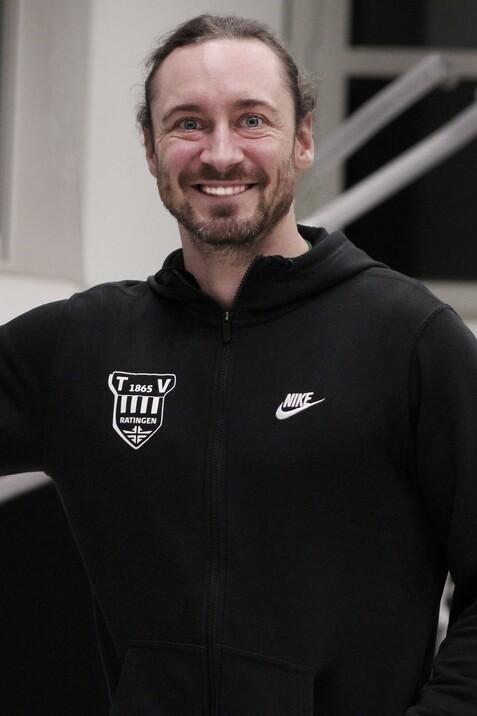 L&auml;chelnder Mann in schwarzer Jacke mit Logo steht in einem Raum mit Fenstern und zeigt mit der Hand auf etwas.