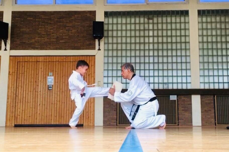 Junge in Karateanzug führt einen Tritt gegen ein Brett, während ein Trainer kniend zusieht. Sportsaal im Hintergrund.