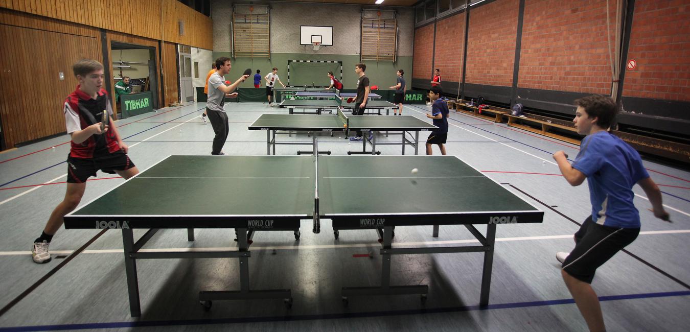 Sportliche Tischtennisspiele in einer Halle mit mehreren Spielern an neun grünen Tischtennisplatten.