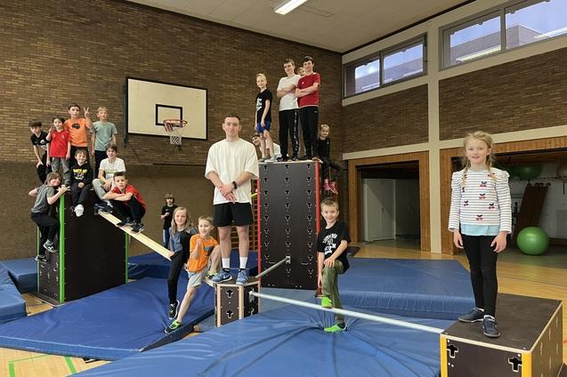 Gruppe von Kindern und Jugendlichen in einer Turnhalle, die auf Kletterger&auml;ten und B&auml;nken spielen und posieren.