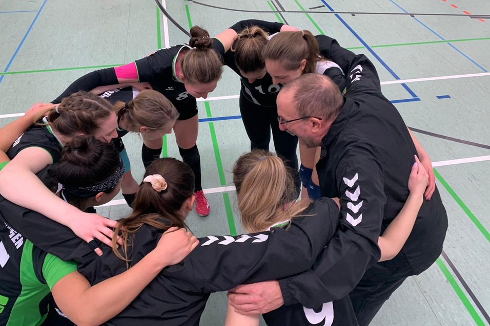 Ein Trainer bespricht sich mit einer Gruppe junger Volleyballspielerinnen im Kreis vor dem Spiel.