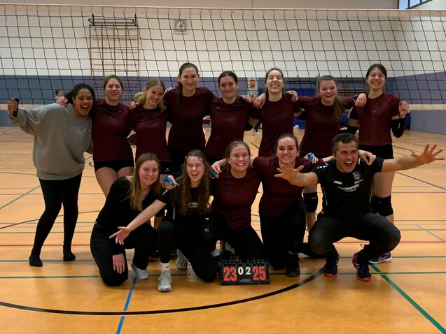 Gruppe von 12 Volleyballspielern in Trainingskleidung posiert freudig vor einem Netz, mit Punktez&auml;hler 23-25 im Vordergrund.