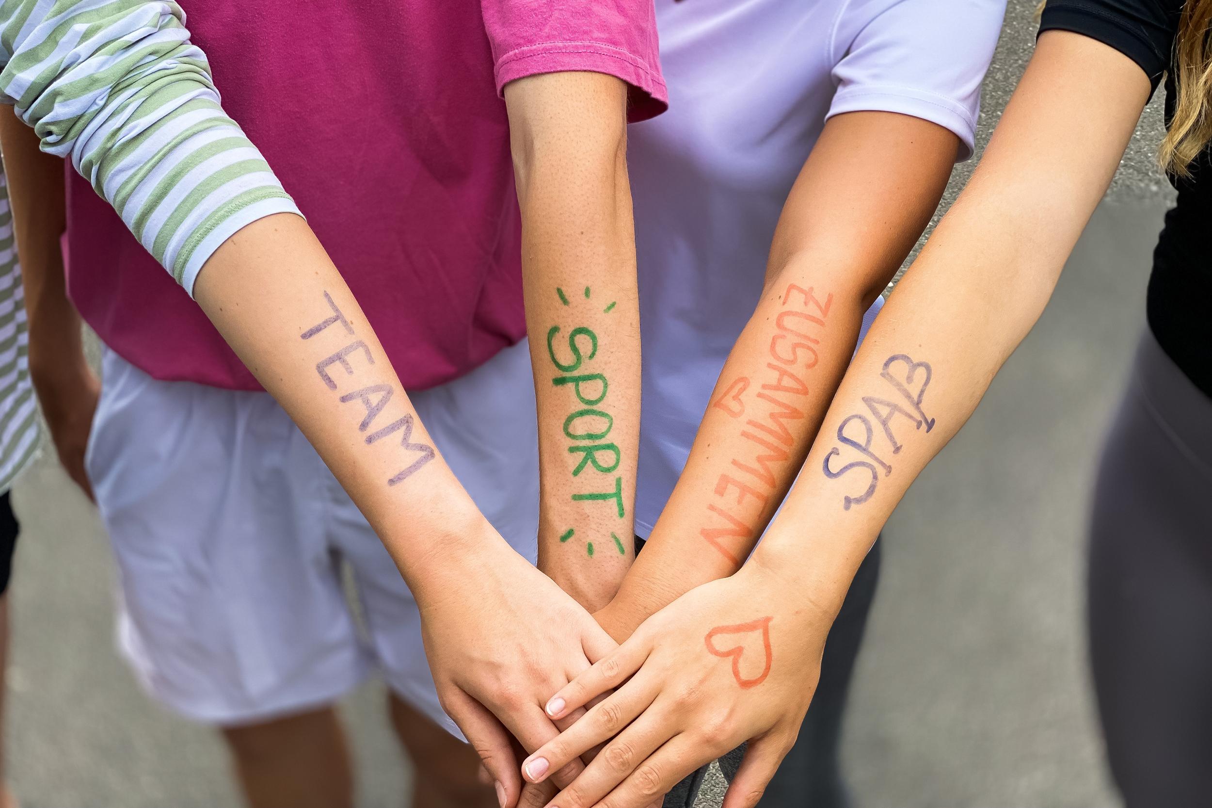 Vier Arme liegen übereinander, mit farbiger Schrift: „TEAM“, „SPORT“, „ZUSAMMEN“, „SPASS“ auf der Haut.