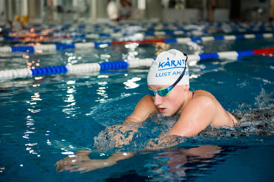 Schwimmerin in blauer Badekappe und Schwimmbrille, die im Wettkampfbecken krault. Spritzer umgeben sie.